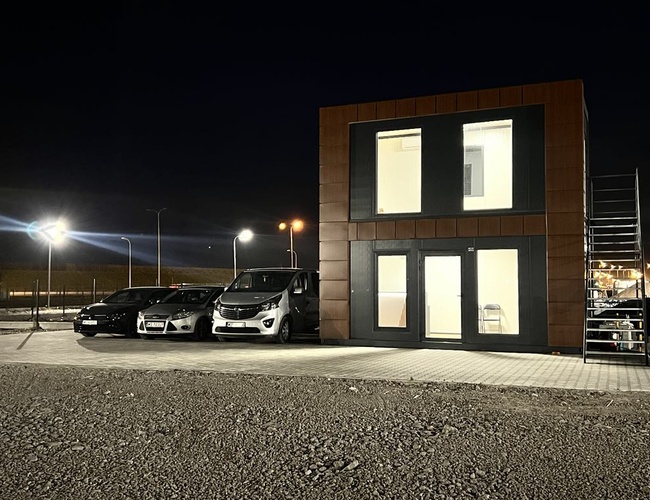 Parkovanie SkyWay - Zadok je dobre osvetlený v noci. Pred budovou stoja zaparkované autá. Pozemok je ohradený.