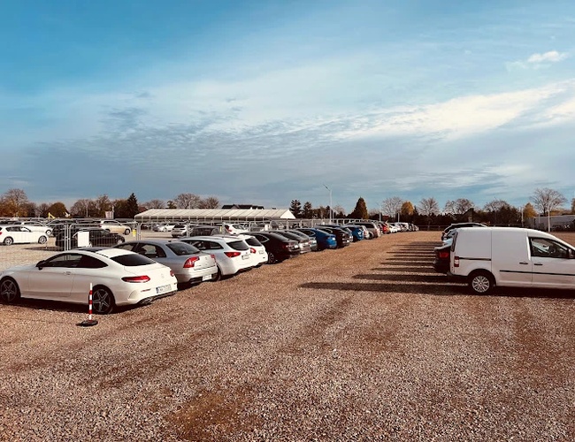 R8 Parkovisko – Na štrkovom dvore sú zaparkované autá. Parkovisko je plné áut rôznych značiek. V pozadí sú vidieť stromy a budova.