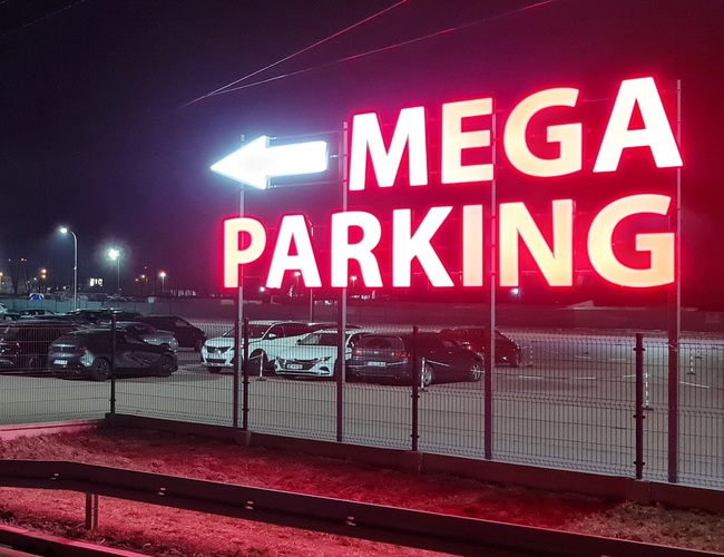 MEGA Parking – un gran letrero iluminado indica la dirección hacia el estacionamiento. El área está cercada y bien iluminada. En el fondo se pueden ver coches estacionados.