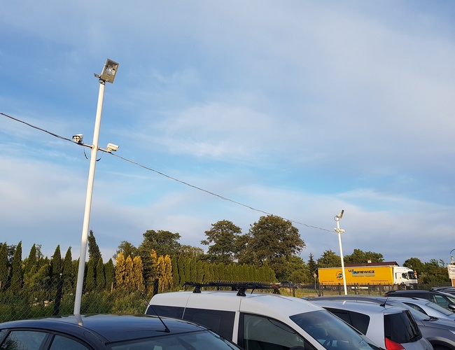 PARKING P24 PREMIUM - Ogrodzony teren z zaparkowanymi samochodami. Na parkingu są latarnie i kamery. W tle widać ciężarówkę za płotem.