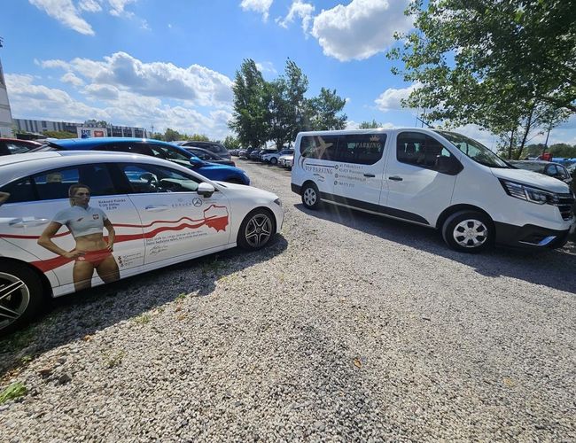 VIP Parking - przy trasie S2. Po żwirowym parkingu stoją dwa pojazdy. Obok drzewa, w tle jest więcej samochodów.