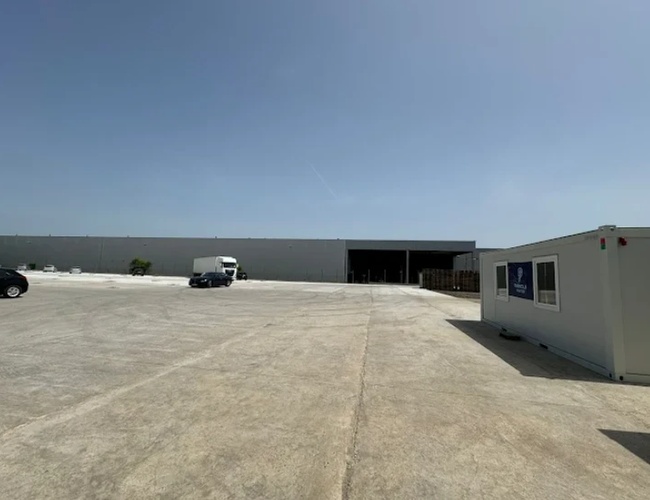 Rozległy, utwardzony plac przemysłowy prowadzący do hali magazynowej, po prawej stronie ustawiony jest biały kontener biurowy, a w oddali widoczne są pojazdy i zabudowania