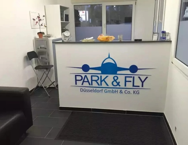 Parking Park&Fly Düsseldorf – Recepcja z logo i ladą. Obok stoi krzesło. W tle widoczne są okna.