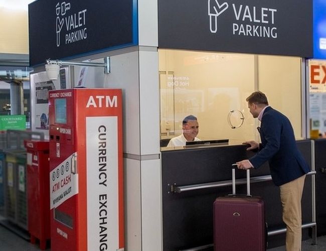 Valet Parking – Lotnisko Chopina - Stanowisko obsługi wewnątrz budynku. Podchodzący mężczyzna z walizką rozmawia z pracownikiem. Obok stoi automat do wymiany walut.