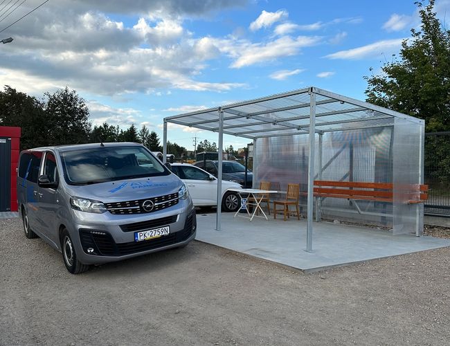 AleParking - Na parkingu zaparkowano szary samochód. Obok znajduje się zadaszone miejsce z ławkami.