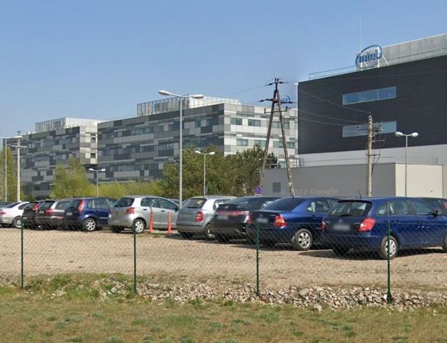Parking Piątka - Parking z ogrodzeniem i stojącymi autami. W tle nowoczesne budynki.