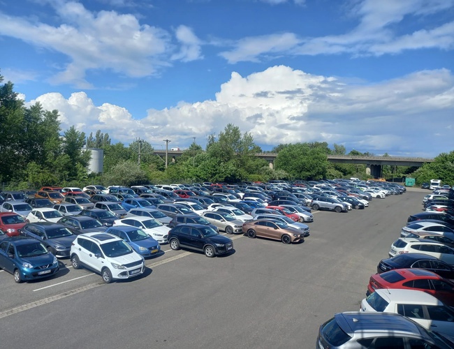 Duży parking pełen samochodów osobowych pod błękitnym niebem z nielicznymi chmurami, w tle drzewa i budynki.