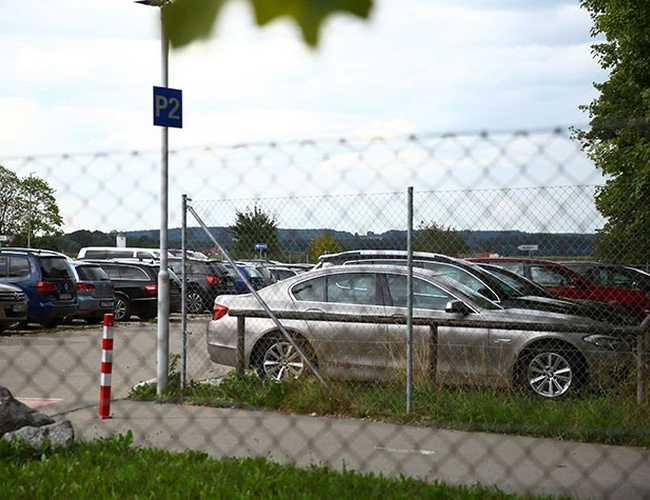 Parking z samochodami widziany przez ogrodzenie z siatki. W tle widoczne są kolejne miejsca postojowe oraz zieleń.