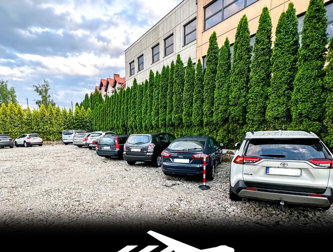 Parksist – Estacionamiento con coches junto al edificio y altos árboles. El terreno es de grava y no está cercado. A su lado hay casas y oficinas.