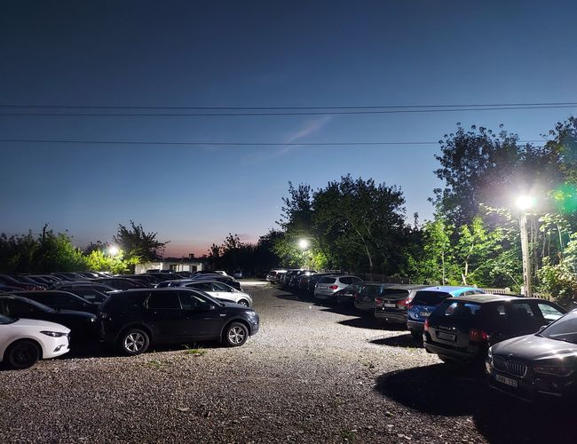 Aparcamiento MPark - En la superficie de grava hay muchos coches. El aparcamiento está iluminado por farolas. Al fondo se ven árboles y arbustos.