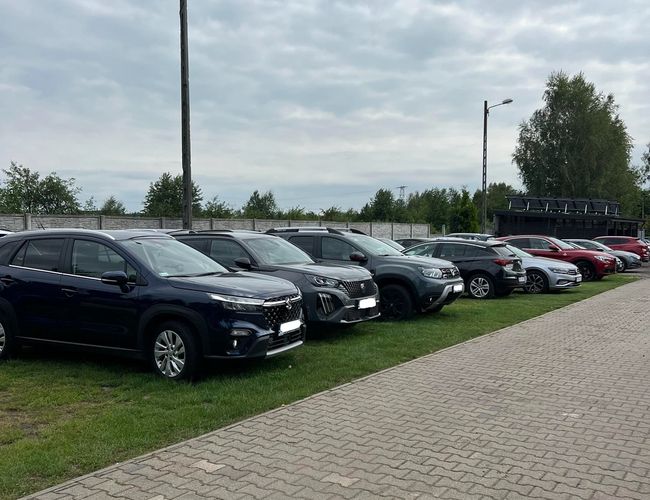 Parking Beta - Coches alineados en fila. El aparcamiento tiene una superficie pavimentada. Se pueden ver farolas y vegetación al fondo.
