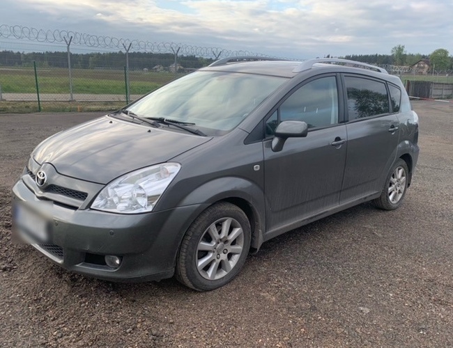 Estacionamiento con vista - Parte frontal de un automóvil gris oscuro sobre una superficie de gravilla. En el fondo se puede ver una cerca de malla y alambre de espino.