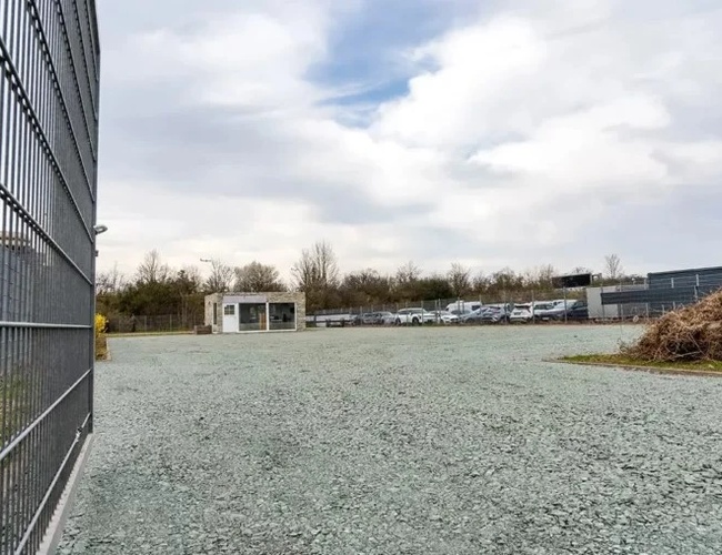 Valet de estacionamiento del transporte – El área está cercada y es espaciosa. Se pueden ver coches aparcados en el estacionamiento y un pequeño edificio.