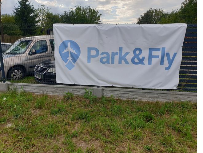 Parkoló Park&Fly – Kerített terület autókkal a háttérben. A kerítésen reklám lóg. Körülötte zöldség van.