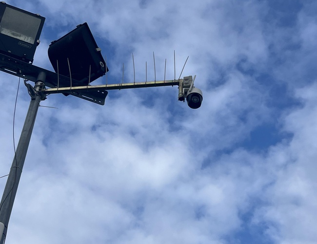 P12 - En el poste están montados reflectores y una cámara. Los reflectores que iluminan durante el día están apagados. En el cielo se ven nubes blancas.