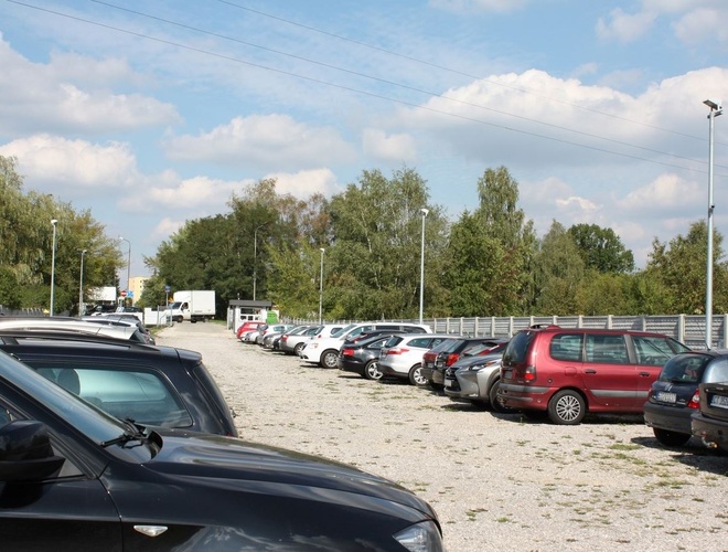 Estacionamiento Pienista 32 - Estacionamiento con numerosos coches. Está rodeado de cercas y árboles. La habitación del conserje se encuentra al fondo.