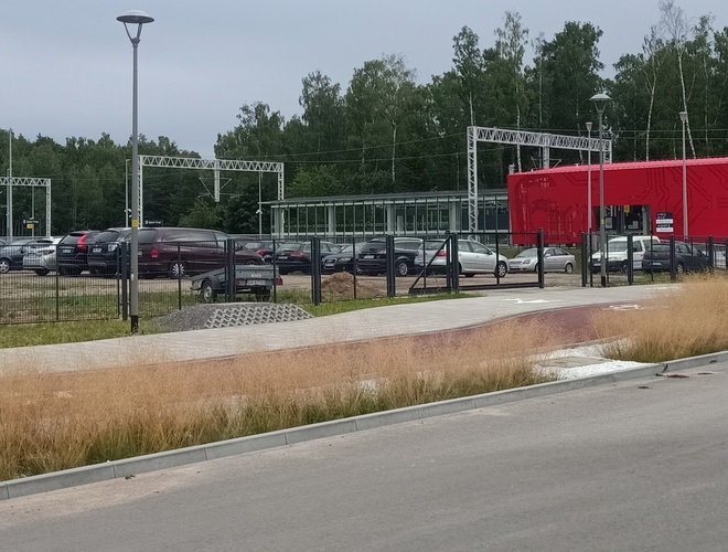 Smart parking - Parking jest ogrodzony metalowym płotem. W tle stoi czerwony budynek z oknami. Obok znajduje się przystanek kolejowy.