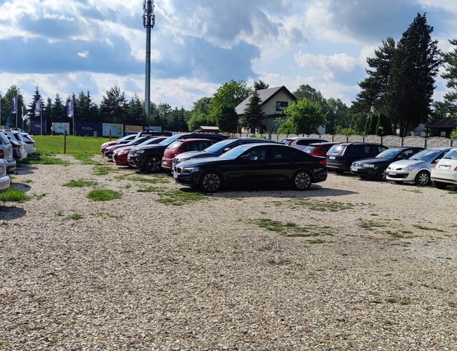 Parking Guardia - Teren żwirowy z zaparkowanymi samochodami. Otaczają go drzewa i ogrodzenie. W tle widoczny jest dom.
