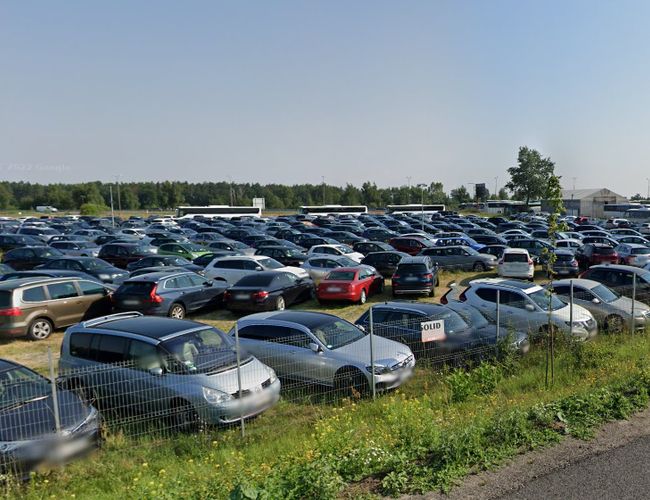 Aparcamiento bajo el molino - hay muchos coches aparcados allí. El terreno está cercado con una valla baja. Al fondo se pueden ver árboles.