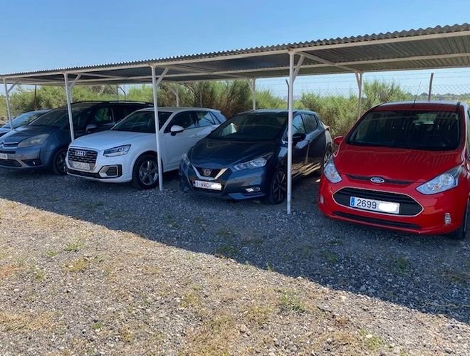 Der beste Parkservice Alicante Überdachte Parkgarage - Autos stehen unter einem Dach. Der Parkplatz ist umzäunt und kiesbedeckt. An den Seiten wachsen Sträucher.