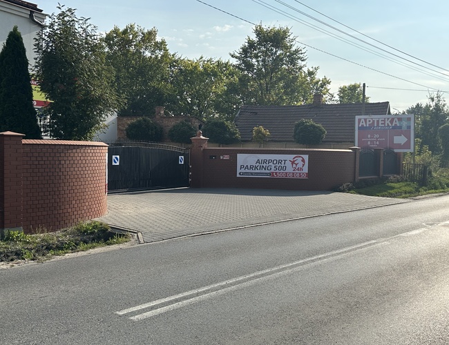 Estacionamiento del Aeropuerto 500 - La entrada al estacionamiento está cercada. Junto a ella hay un cartel que indica dirección a la farmacia. La zona está cubierta de pavimento.
