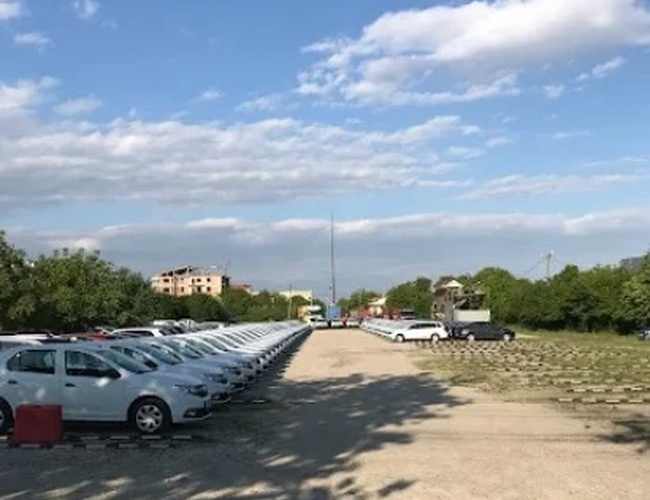 Widok żwirowego parkingu z wieloma zaparkowanymi samochodami, drzewa po bokach i niebo z chmurami.