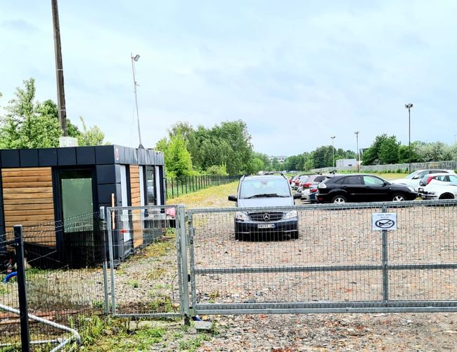 Krispit Parking 24 – parking ogrodzony z bramą wjazdową. Widoczne pojazdy zaparkowane na żwirowym podłożu. Obok stoi budka dozorcy.