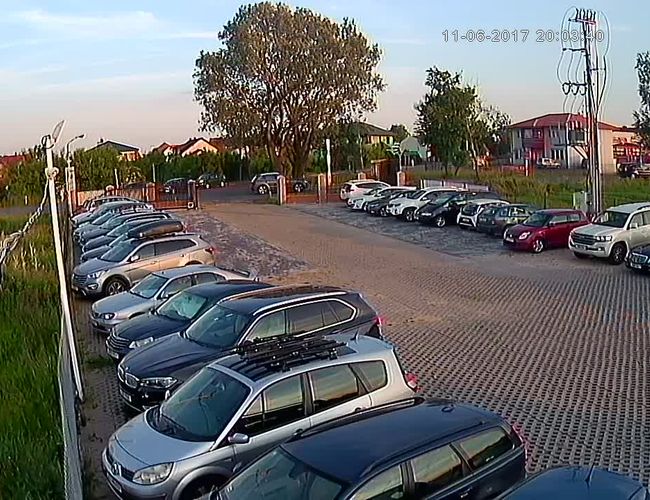PARKING P24 PREMIUM – Na parkingu zaparkowane są samochody w dwóch rzędach. Teren jest otoczony ogrodzeniem i ma trawiaste otoczenie. W tle widać drzewa i budynki.