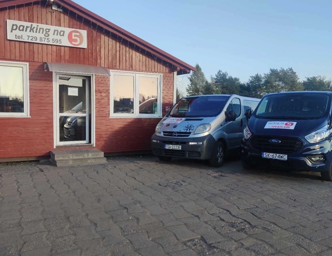 Parking na 5 – Przed budynkiem stoją dwa samochody. Budynek jest drewniany z czerwonymi ścianami. Obok jest utwardzony plac.