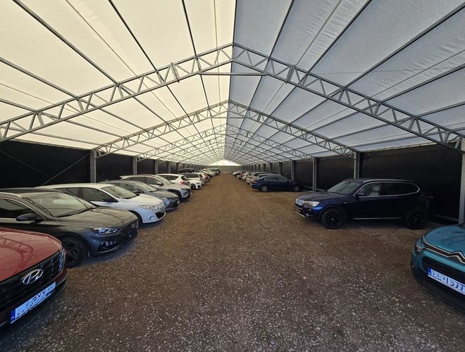 AleParking zadaszony - na parkingu stoją zaparkowane samochody. Teren jest kryty dachem. Samochody są ustawione w równych rzędach.