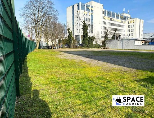 Паркування на космічній стоянці – територія огороджена металевим парканом. Біля газону видніються будівлі. На фоні світлий, сучасний житловий комплекс.