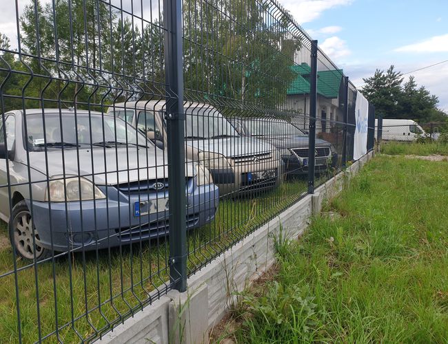 Parking Park&Fly - na zdjęciu kilka samochodów za ogrodzeniem. Teren jest otoczony siatką i porośnięty trawą. W tle znajdują się drzewa.