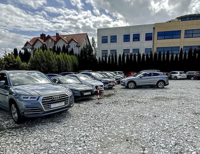 Parksist - Parkovisko s kamienistým podložím, plné zaparkovaných áut. Na pozadí stoja budovy a stromy.