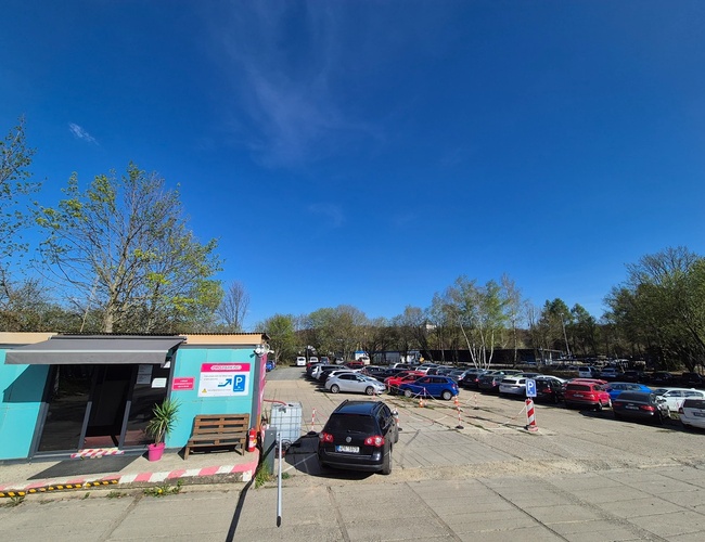 PRG Parking - Parkovisko je otvorené a priestranné. Je tu zaparkovaných mnoho áut. Vľavo je vidieť modrú budovu.