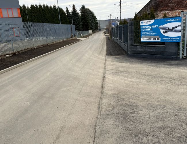 FlyParking – Ogrodzony teren z bramą i reklamą usług. Droga prowadzi do wejścia, otoczona drzewami i płotem.