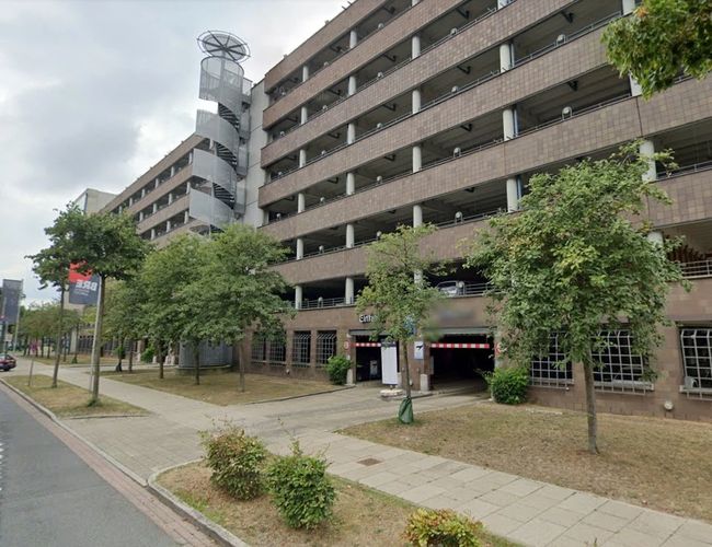 Widok na wielopoziomowy parking od strony ulicy. Przy budynku znajdują się chodnik, drzewa oraz wejście i wjazd na parking.