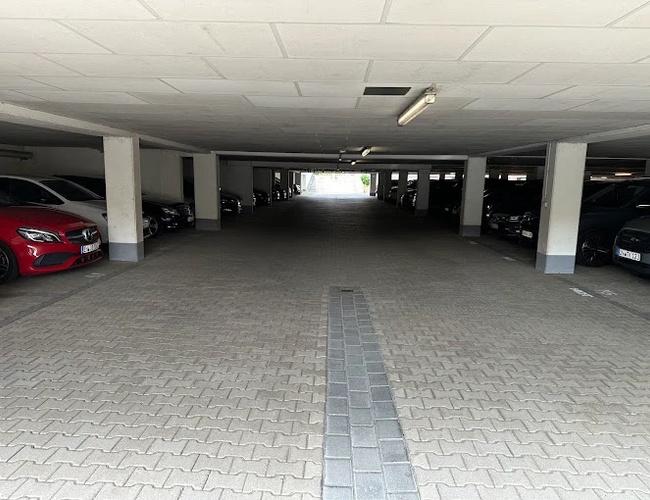 Parking Marspark Covered Valet - Zadaszony parking z zaparkowanymi samochodami. Betonowa nawierzchnia i kolumny podtrzymujące sufit. Oświetlenie jarzeniowe nad drogą.