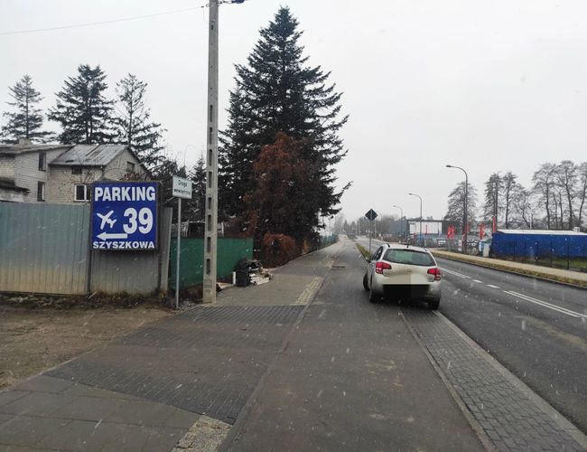 Parking Szyszkowa 39 - Teren nieoświetlony, ogrodzony. Tablica z nazwą parkingu przy drodze. Obok znajduje się zaparkowany samochód.
