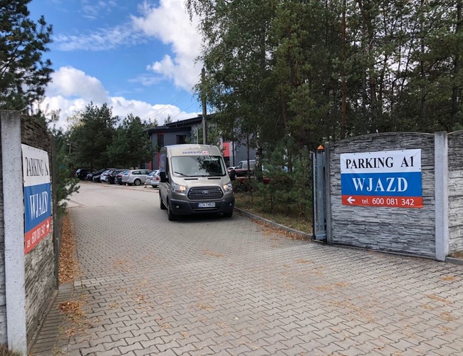 Parking A1 – Wjazd jest szeroki, otoczony drzewami. Widoczny jest bus i zaparkowane samochody. Teren wydaje się przestronny.