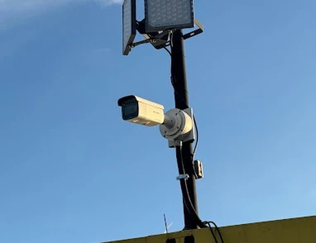 Estacionamiento Parktiger Transfer - Cámara de vigilancia fijada a un alto poste. Al lado de la cámara hay una luz instalada. La cámara está orientada hacia abajo.