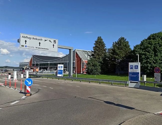 Widok na wjazd na teren obiektu z drogą dojazdową, oznakowaniem parkingu oraz budynkiem w tle.