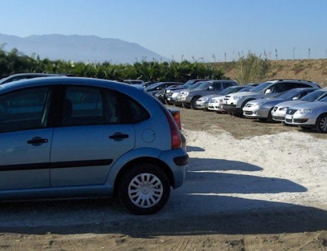 Parking PedroCar – Na zdjęciu widać otwarty plac. Kilka samochodów jest zaparkowanych na żwirowej powierzchni. W tle znajdują się drzewa i góry.
