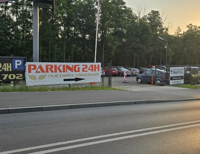 Victory Cars - Parking 24h znajduje się przy ruchliwej drodze. Jest ogrodzony i dobrze oświetlony. Obok parkuje kilka samochodów.