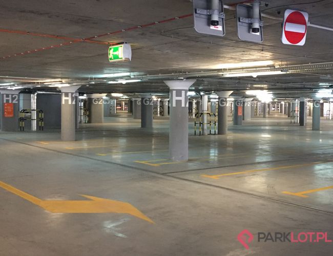 Oficjalny Parking Lotniska P2 - Podziemny parking z oznakowaniem sektorów. Znajduje się oświetlenie oraz znaki ewakuacyjne. Widać szerokie alejki.