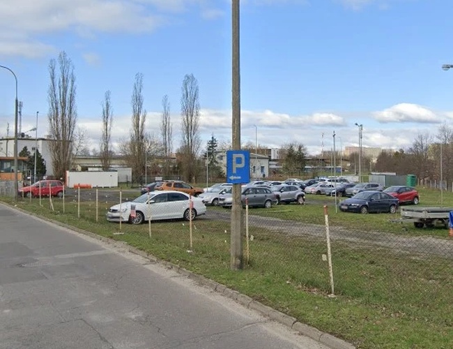 Odlotowy Parking BZG - Na trawie stoją zaparkowane samochody. Teren jest ogrodzony siatką. W tle widać drzewa i budynki.