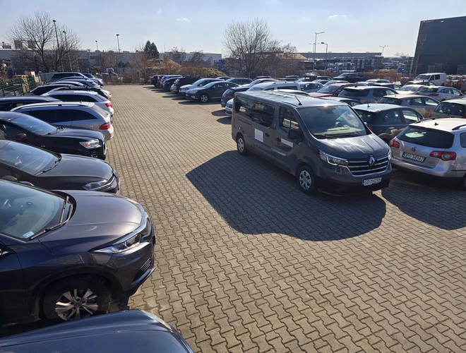 Parking Olo Car Park - Teren pełen zaparkowanych samochodów. Powierzchnia jest wyłożona kostką brukową. Otaczają go ogrodzenia i drzewa.