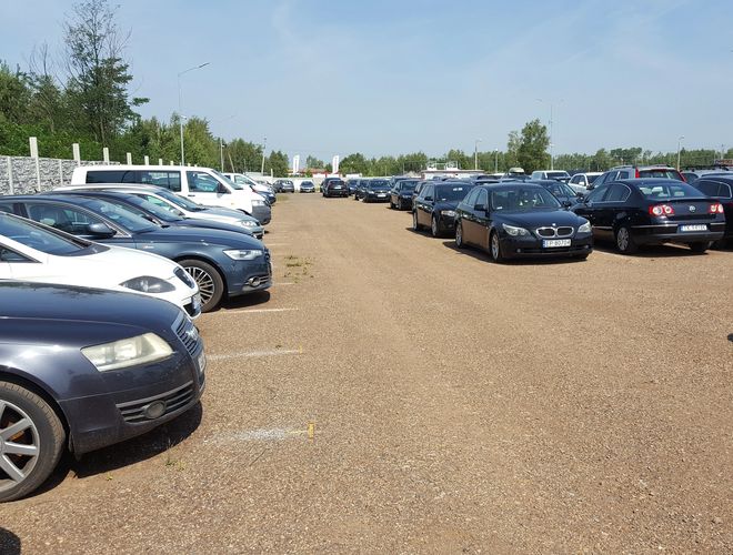 AAP-66 – Parking z licznymi samochodami osobowymi. Teren jest nieogrodzony i żwirowy. W tle widoczna zieleń i latarnie.