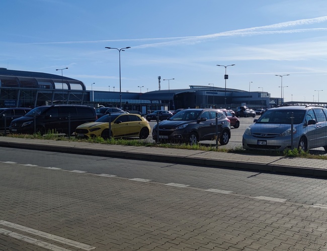 Oficiálne parkovisko P4 – Parkovisko je ohradené a osvetlené. Automobily stoja v radoch. Na pozadí je vidieť budovu letiska.