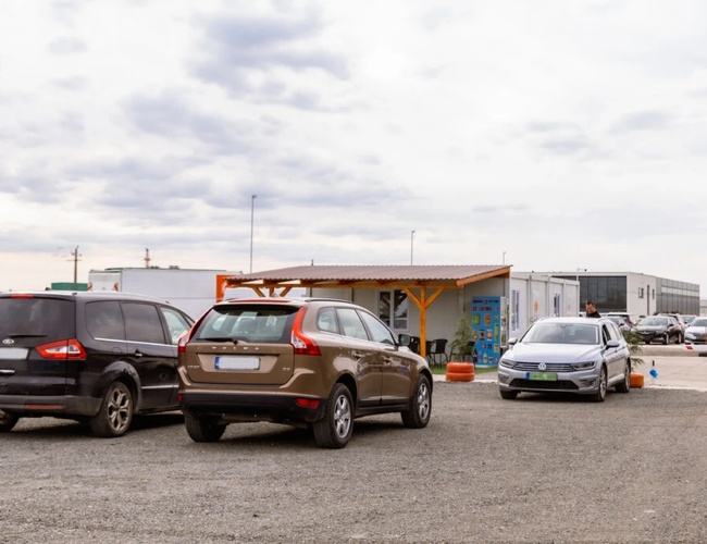 Automóviles visibles estacionados en el aparcamiento de superficie dura. Al fondo, un edificio y más automóviles.