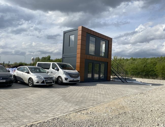 SkyWay Parkplatz – Parkplatz mit mehreren Autos neben dem Gebäude. Das Gelände ist eingezäunt und mit Pflastersteinen befestigt. In der Ferne sind Bäume zu sehen.